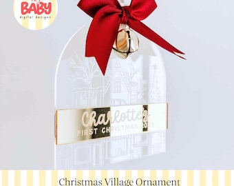 Könnte beinhalten: Ein bogenförmiger, transparenter Acryl-Anhänger mit einer weiß gravierten Weihnachtsdorf-Szene. Ein goldener, spiegelnder horizontaler Streifen zeigt den Text Charlotte's FIRST CHRISTMAS 2025. Eine rote Satinschleife und ein goldenes Glöckchen sind befestigt.