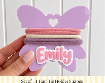 Könnte beinhalten: Ein hellvioletter, schmetterlingsförmiger Haargummihalter aus Acryl mit einem herzförmigen Ausschnitt oben und dem Namen Emily in erhabener rosa Schrift. Drei Haargummis in Altrosa, Creme und Mauve sind um die Mitte gewickelt.