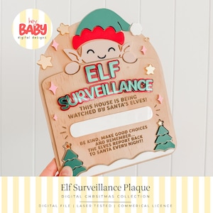 May include: A wooden arch-shaped plaque featuring a cartoon elf at the top and the text ELF SURVEILLANCE, THIS HOUSE IS BEING WATCHED BY SANTA'S ELVES, and BE KIND, MAKE GOOD CHOICES AND REMEMBER THE ELVES REPORT BACK TO SANTA EVERY NIGHT.