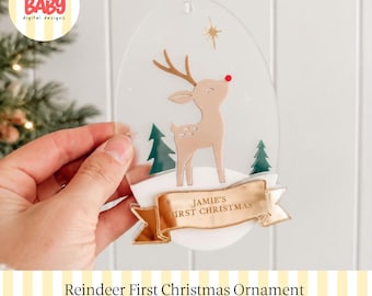 Könnte beinhalten: Ein durchsichtiger, ovaler Anhänger mit einem beigen Rentier mit roter Nase, zwei grünen Tannenbäumen und einem goldenen Banner unten mit dem Text JAMIE'S FIRST CHRISTMAS. Das Design enthält einen kleinen goldenen Stern und weiße Schneeelemente.