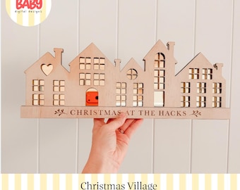 Könnte beinhalten: Eine hellfarbene, lasergeschnittene Weihnachtsdorf-Silhouette aus Holz mit mehreren Hausstrukturen, quadratischen Fenstern, einem herzförmigen Ausschnitt und einer roten Tür. Die Basis ist mit dem Text CHRISTMAS AT THE HACKS graviert.