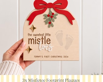 Könnte beinhalten: Ein gewölbtes Holzschild mit einer roten Schleife und Mistelzweigen oben, zwei goldenen Sternen und dem Text "the sweetest little mistletoes" sowie zwei Babyfußabdrücken. Der untere Text lautet "TOMMY'S FIRST CHRISTMAS 2026".
