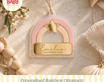 Könnte beinhalten: Ein bogenförmiger Anhänger aus mattiertem Acryl mit einem rosa und pfirsichfarbenen Regenbogen, einem goldenen Stern und einer goldenen rechteckigen Plakette mit der Gravur Emelia und dem Text CHRISTMAS 2026, an einer dünnen Schnur.