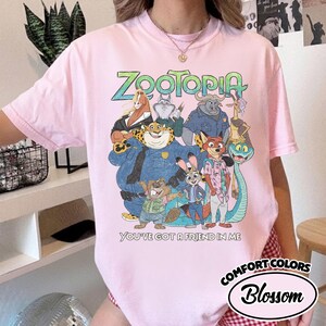 May include: Pink t-shirt featuring a colorful graphic of characters from the animated movie "Zootopia." The design includes the movie title and the phrase "You've Got a Friend in Me." The shirt is a Comfort Colors brand.