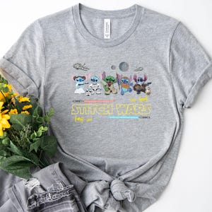 May include: A gray t-shirt featuring a graphic of several cartoon characters in a Star Wars theme. The text "Stitch Wars" is printed in yellow with blue and red lightsabers. The shirt is made of a soft material.