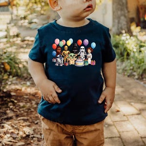 Puede incluir: Una camiseta azul marino con un gr&aacute;fico colorido de personajes de Star Wars, globos y una tarta de cumplea&ntilde;os. El dise&ntilde;o incluye personajes como R2-D2, C-3PO, Darth Vader y Baby Yoda. La camiseta la lleva un ni&ntilde;o.