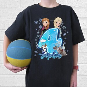 Pode incluir: T-shirt preto com um gr&aacute;fico colorido de Anna, Elsa, Olaf e Sven de Frozen, com o n&uacute;mero 4 e o nome "Sophia". O design inclui flocos de neve. A pessoa est&aacute; vestindo a camiseta e segurando uma bola azul e amarela.
