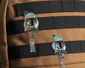 May include: Two Boba Fett badge reels with a holographic design, attached to a brown backpack with black straps. The reels feature a cartoon version of the character, with a green helmet and red accents.