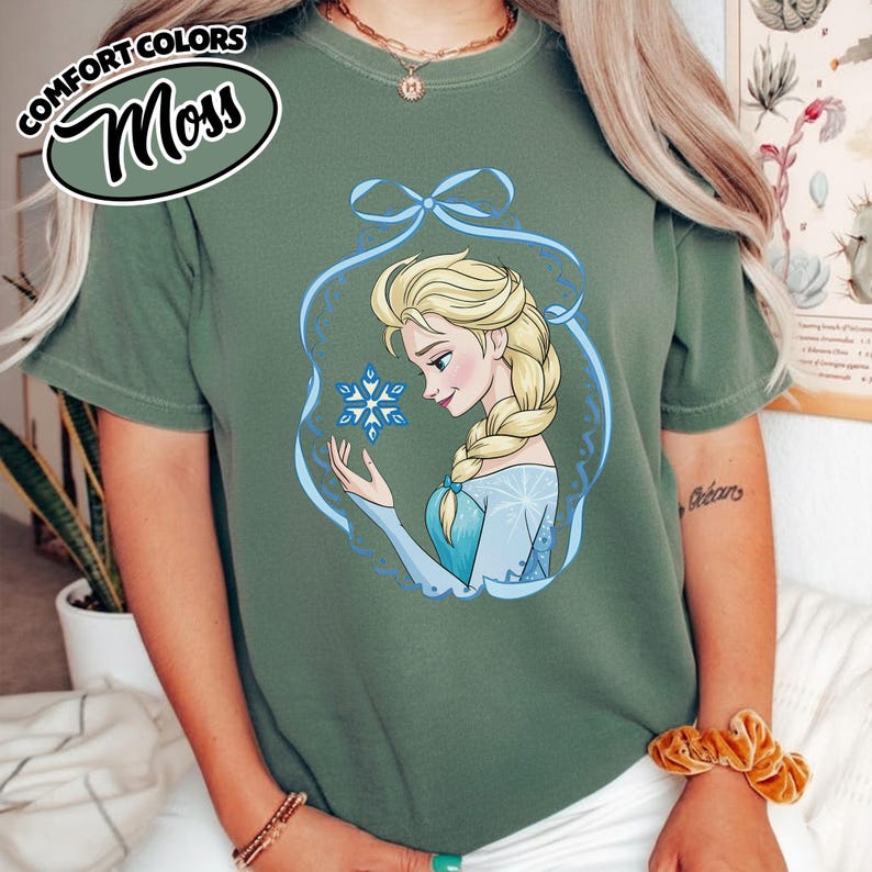 May include: Moss green t-shirt featuring a graphic of a blonde woman with a blue snowflake. The woman is wearing a blue dress and is framed by a blue decorative border. The words "COMFORT COLORS" and "Moss" are in a circular design.