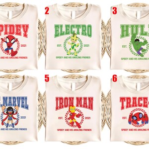May include: Cream-colored t-shirts featuring superhero designs. Each shirt displays a different character's name in bold letters, such as "Spidey," "Electro," and "Hulk," along with the text "Est. 2021" and the phrase "Spidey and his Amazing Friends."