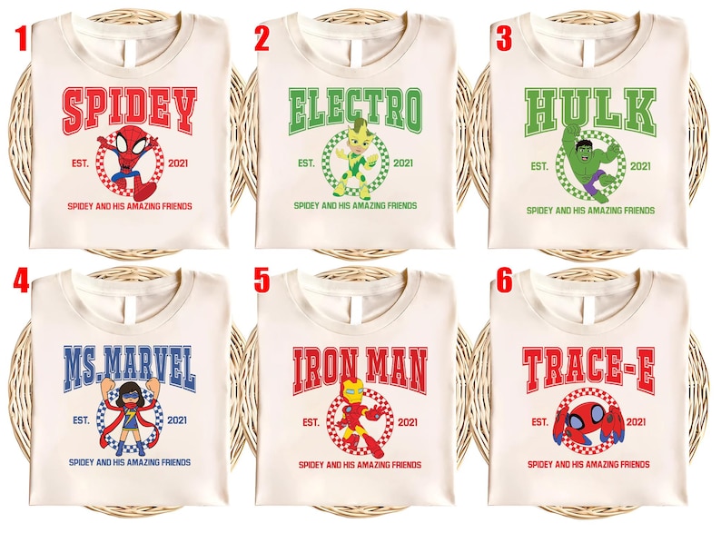 May include: Cream-colored t-shirts featuring superhero designs. Each shirt displays a different character's name in bold letters, such as "Spidey," "Electro," and "Hulk," along with the text "Est. 2021" and the phrase "Spidey and his Amazing Friends."