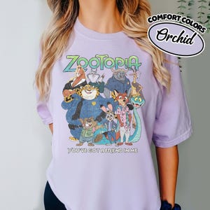 May include: Orchid-colored t-shirt featuring a colorful graphic of characters from the animated film Zootopia. The design includes the film's title and the phrase "You've Got a Friend in Me". The shirt is made of Comfort Colors material.