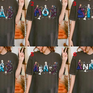 Puede incluir: Camisetas de color gris oscuro con varios nombres en una fuente colorida y estilizada, cada una con personajes de una popular pel&iacute;cula de animaci&oacute;n. Los nombres incluyen "DAD", "MOM", "SIS", "BRO", "HENRY" y "ANNA".