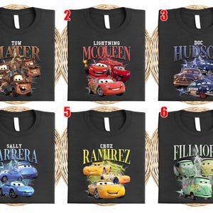 May include: Six black t-shirts featuring characters from the film "Cars." Each shirt displays a different character's name and image: Mater, Lightning McQueen, Doc Hudson, Sally Carrera, Cruz Ramirez, and Fillmore.