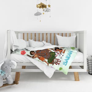 K&ouml;nnte beinhalten: Eine wei&szlig;e Babydecke mit einer bunten Cartoon-Illustration eines M&auml;dchens mit langen braunen Haaren, einem Schwein und einem Hahn, wobei der Name SARAH in blauen Buchstaben aufgedruckt ist. Die Decke liegt auf einem wei&szlig;en Holzkinderbett.