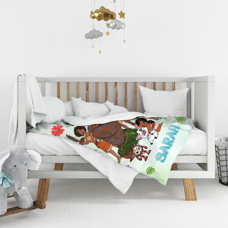 K&ouml;nnte beinhalten: Eine wei&szlig;e Babydecke mit einer bunten Cartoon-Illustration eines M&auml;dchens mit langen braunen Haaren, einem Schwein und einem Hahn, wobei der Name SARAH in blauen Buchstaben aufgedruckt ist. Die Decke liegt auf einem wei&szlig;en Holzkinderbett.
