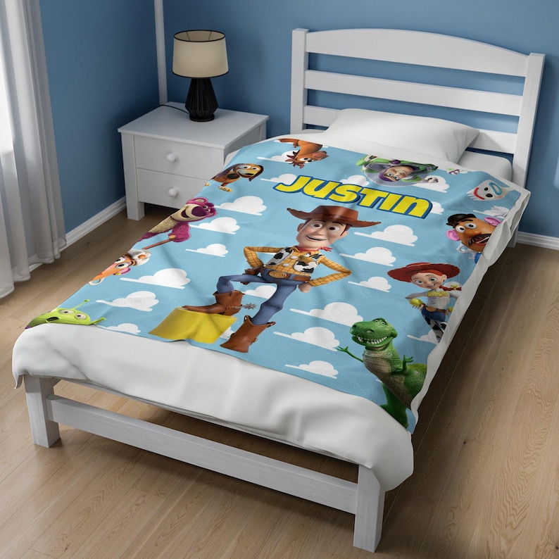 K&ouml;nnte beinhalten: Eine hellblaue Fleecedecke mit einem wiederkehrenden Muster aus Toy Story-Figuren wie Woody, Buzz Lightyear, Rex und Mr. Potato Head vor einem wei&szlig;en Wolkenhintergrund. Der Name JUSTIN ist in gro&szlig;en gelben Buchstaben oben aufgedruckt.