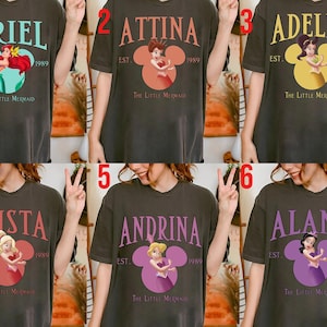 Puede incluir: Camisetas de color gris oscuro con personajes de "La Sirenita". Cada camiseta muestra un nombre en una fuente estilizada sobre un gr&aacute;fico en forma de Mickey Mouse con una ilustraci&oacute;n de personaje y el texto "EST. 1989 The Little Mermaid."