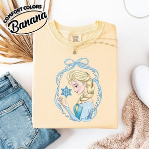 May include: A light yellow sweatshirt with a graphic of a cartoon woman with blonde hair and a blue dress holding a snowflake. The sweatshirt has the text "COMFORT COLORS" and "Banana" in a black oval.