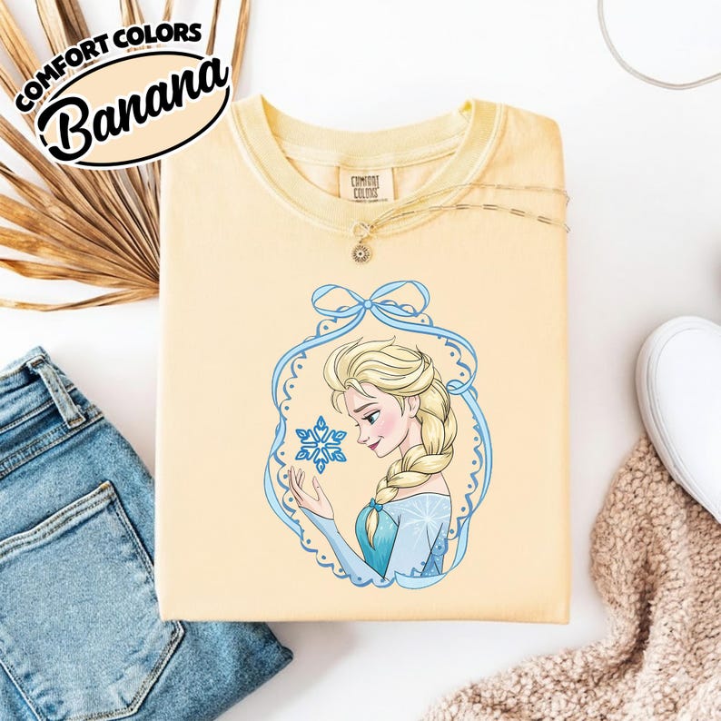 May include: A light yellow sweatshirt with a graphic of a cartoon woman with blonde hair and a blue dress holding a snowflake. The sweatshirt has the text "COMFORT COLORS" and "Banana" in a black oval.