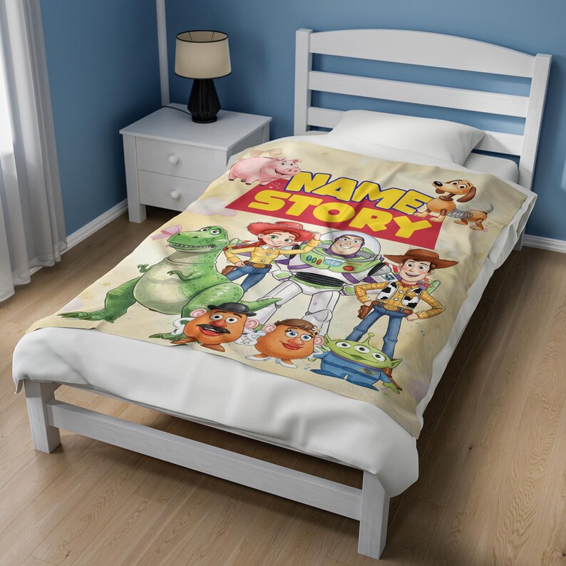 May include: A cream-colored fleece blanket featuring a colorful graphic print of various animated characters, including a green dinosaur, a space ranger, a cowboy, and a cowgirl, with the text NAME STORY printed in bold red and yellow letters.