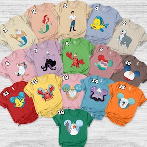 May include: A collection of short-sleeved t-shirts in various colors, each featuring a cartoon character design. Designs include a mermaid, a crab, a dog, and other characters. The shirts are arranged on a light wood surface.