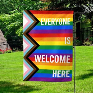 May include: A colorful garden flag with the text "EVERYONE IS WELCOME HERE." The flag features a rainbow design with a progressive pride flag on the left side. The flag is hanging on a black metal stand in a grassy yard.