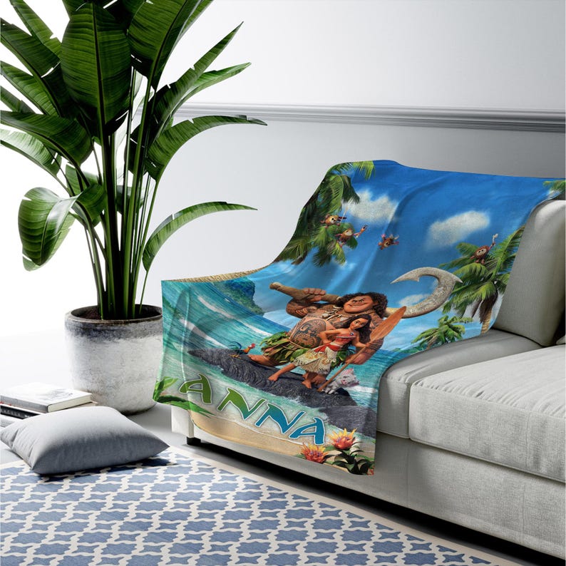 Op de afbeelding: Een zachte plaid met een levendige tropische sc&egrave;ne met de personages Maui en Moana die op een rots staan, omringd door palmbomen en oceaangolven. De naam ANNA is in grote, dikke, lichtblauwe letters onderaan het ontwerp gedrukt.