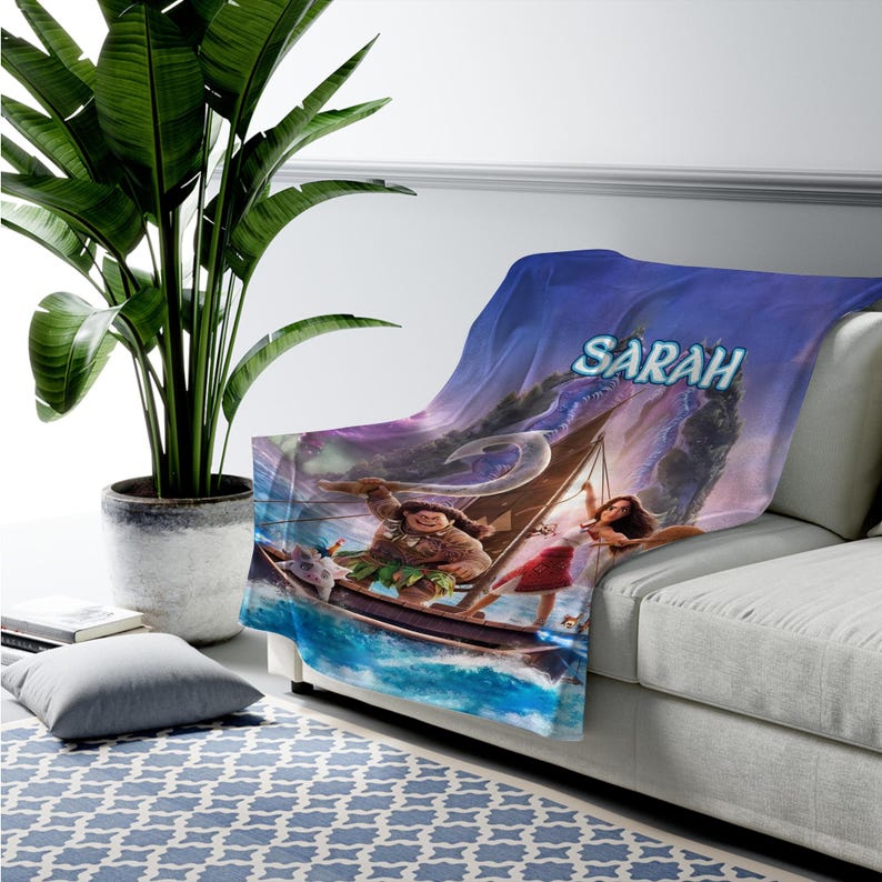 May include: A soft fleece throw blanket featuring a vibrant, full-color graphic print of the characters Maui and Moana sailing on a wooden boat across blue ocean waves. The name SARAH is printed in large, light blue letters at the top center.