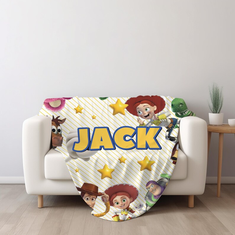 May include: A white throw blanket featuring a yellow diagonal striped pattern, yellow stars, and various animated characters including a cowboy, a cowgirl, and a space ranger. The name JACK is printed in large, bold, yellow letters across the center.