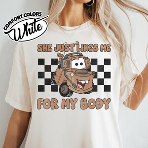 May include: A cream-colored short-sleeve t-shirt featuring a graphic of a brown tow truck character wearing headphones. Behind the truck is a black and white checkered pattern. The text reads "SHE JUST LIKES ME FOR MY BODY" in brown letters.