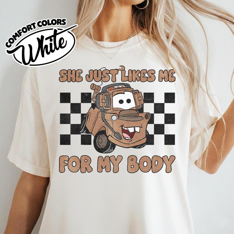 May include: A cream-colored short-sleeve t-shirt featuring a graphic of a brown tow truck character wearing headphones. Behind the truck is a black and white checkered pattern. The text reads "SHE JUST LIKES ME FOR MY BODY" in brown letters.