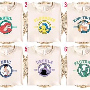 Puede incluir: Seis camisetas color crema con dise&ntilde;os de personajes de La Sirenita. Cada camiseta presenta el nombre de un personaje en un c&iacute;rculo con "EST. 1989". Los personajes incluyen Ariel, Flounder, Rey Trit&oacute;n, Eric, &Uacute;rsula y Flotsam.