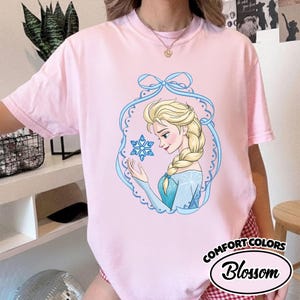May include: A light pink Comfort Colors t-shirt featuring a cartoon illustration of a woman with blonde hair and a blue dress holding a snowflake. The illustration is framed by a blue decorative border. The text "Comfort Colors Blossom" is printed on the bottom right.