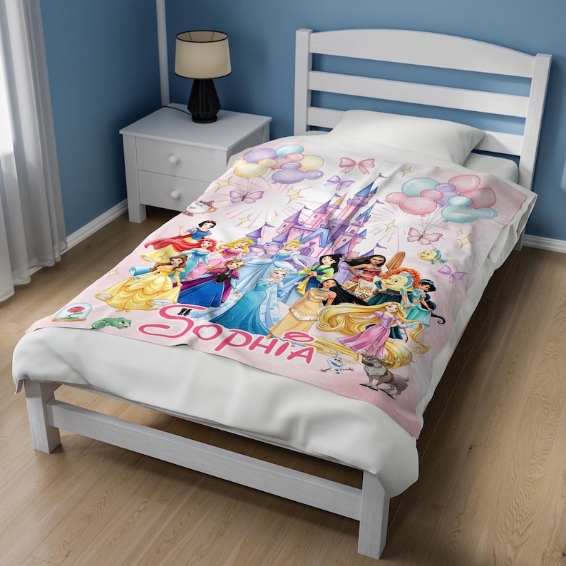 May include: A white blanket featuring a colourful print of various animated princesses, a castle, balloons, and butterflies. The name Sophia is printed in large pink cursive letters at the bottom. The blanket is spread across a white bed frame.