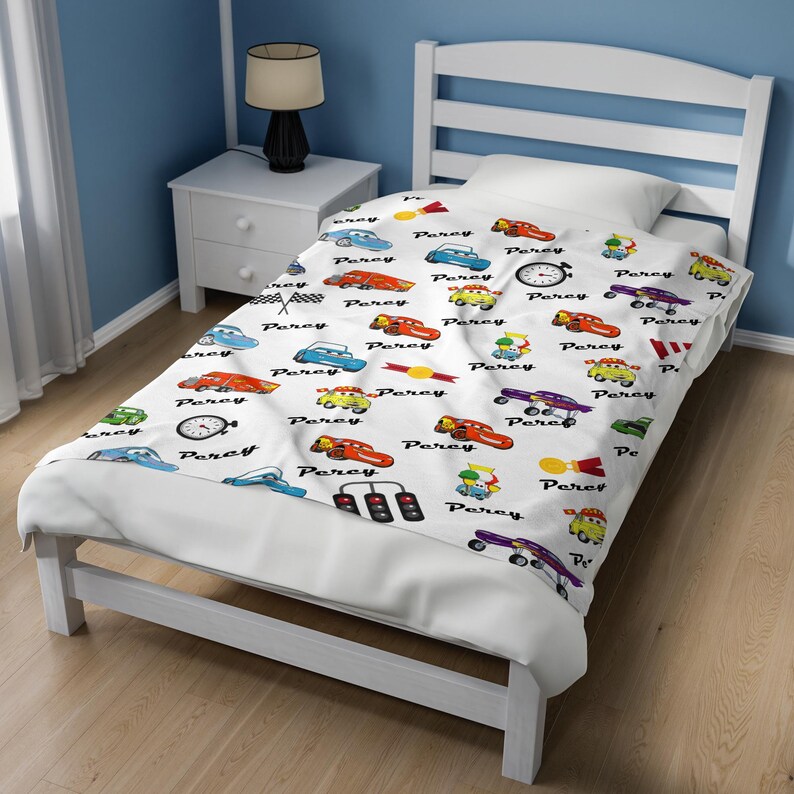 May include: A white blanket featuring a repeating pattern of colorful cartoon race cars, stoplights, checkered flags, and stopwatches, interspersed with the name Percy printed in a black script font across the fabric surface.