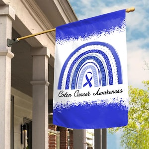 May include: A white and blue flag with a rainbow design and the words "Colon Cancer Awareness." The flag features a blue ribbon, a symbol of colon cancer awareness. The flag is hanging outside on a porch.