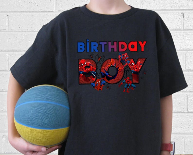 Może przedstawiać: Czarna koszulka z napisem "BIRTHDAY BOY" w kolorze niebieskim i czerwonym, z grafiką Spider-Mana. Koszulka jest trzymana przez osobę, widoczna jest niebiesko-ż&oacute;łta piłka do koszyk&oacute;wki.
