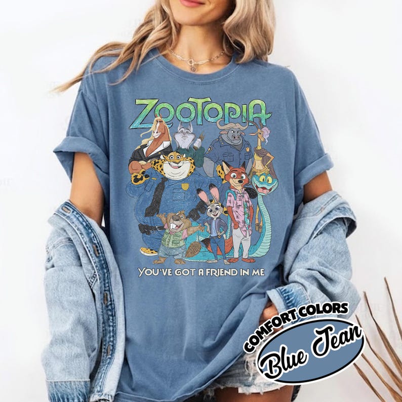 May include: Blue graphic tee featuring characters from the animated film Zootopia. The shirt displays the word "Zootopia" in green and blue, along with the phrase "You've got a friend in me". The shirt is a Comfort Colors Blue Jean.