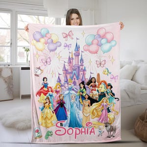 May include: A light pink fleece blanket featuring a large purple and blue castle, colourful balloons, butterflies, and a group of animated princesses. The name Sophia is printed in large pink cursive letters at the bottom with various sidekick characters.