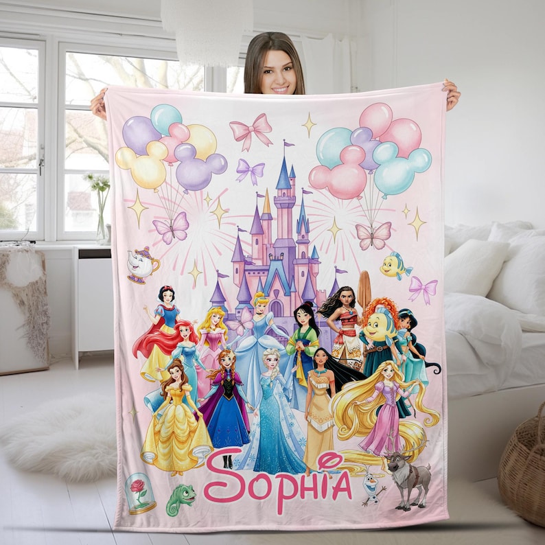 May include: A light pink fleece blanket featuring a large purple and blue castle, colourful balloons, butterflies, and a group of animated princesses. The name Sophia is printed in large pink cursive letters at the bottom with various sidekick characters.
