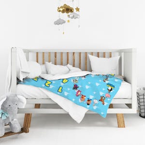 May include: A light blue baby blanket featuring a repeating pattern of cartoon characters and yellow numbers against a white cloud background. The blanket is draped over a white wooden crib with vertical slats and a white mattress inside.