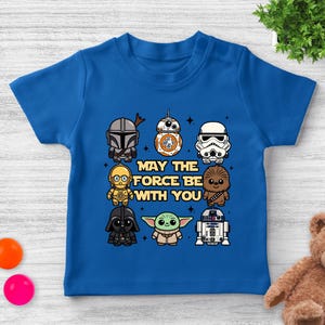 May include: A blue short-sleeved t-shirt featuring cartoon illustrations of Star Wars characters, including Darth Vader, Yoda, and R2-D2. The text "MAY THE FORCE BE WITH YOU" is printed in the center of the shirt.