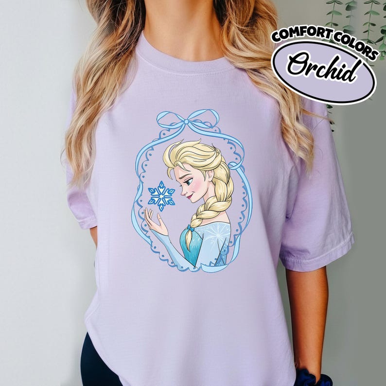 May include: A light purple t-shirt featuring a cartoon illustration of a woman with blonde hair in a braid, wearing a blue dress, and holding a snowflake. The shirt has the words "COMFORT COLORS" and "Orchid" printed on it.