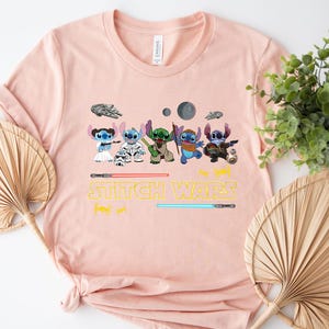May include: A light pink t-shirt featuring a graphic of several Stitch characters in Star Wars-themed costumes. The text "Stitch Wars" is printed in yellow below the characters, with lightsabers and space-themed illustrations.