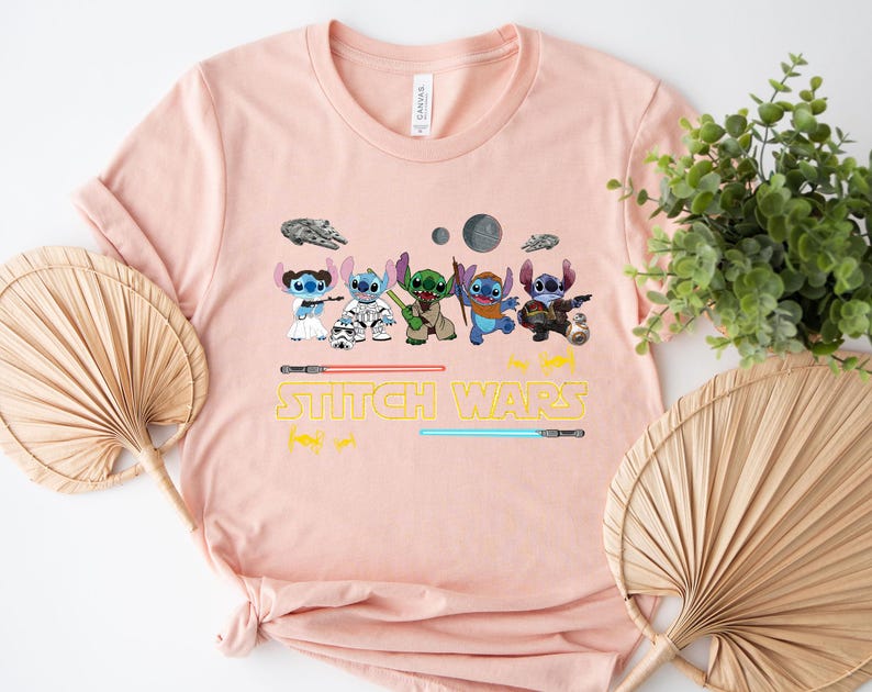 May include: A light pink t-shirt featuring a graphic of several Stitch characters in Star Wars-themed costumes. The text "Stitch Wars" is printed in yellow below the characters, with lightsabers and space-themed illustrations.