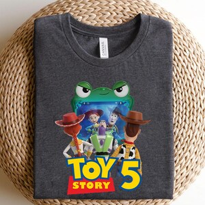 May include: A dark heather gray t-shirt featuring a graphic of the Toy Story characters Jessie, Buzz Lightyear, and Woody looking at a green frog-shaped screen, with the text Toy Story 5 printed in large yellow and red letters below the characters.