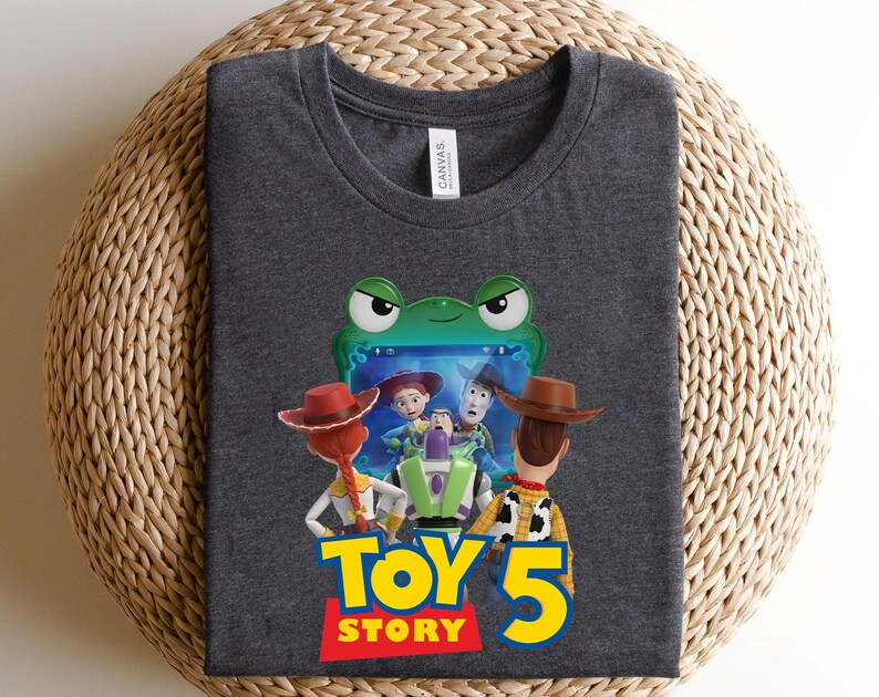 May include: A dark heather gray t-shirt featuring a graphic of the Toy Story characters Jessie, Buzz Lightyear, and Woody looking at a green frog-shaped screen, with the text Toy Story 5 printed in large yellow and red letters below the characters.