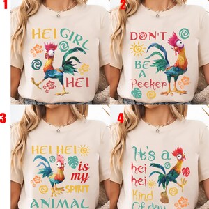 Op de afbeelding: Vier beige t-shirts met kleurrijke haan-afbeeldingen en tekst. Het eerste shirt zegt "Hei Girl Hei", het tweede zegt "Don't Be a Peeker", het derde zegt "Hei Hei is my Spirit Animal" en het vierde zegt "It's a Hei Hei Kind of Day".