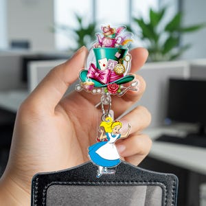 May include: An Alice in Wonderland themed badge reel featuring a green top hat, pink bow, and a cartoon image of Alice. The reel also includes a pocket for an ID card.